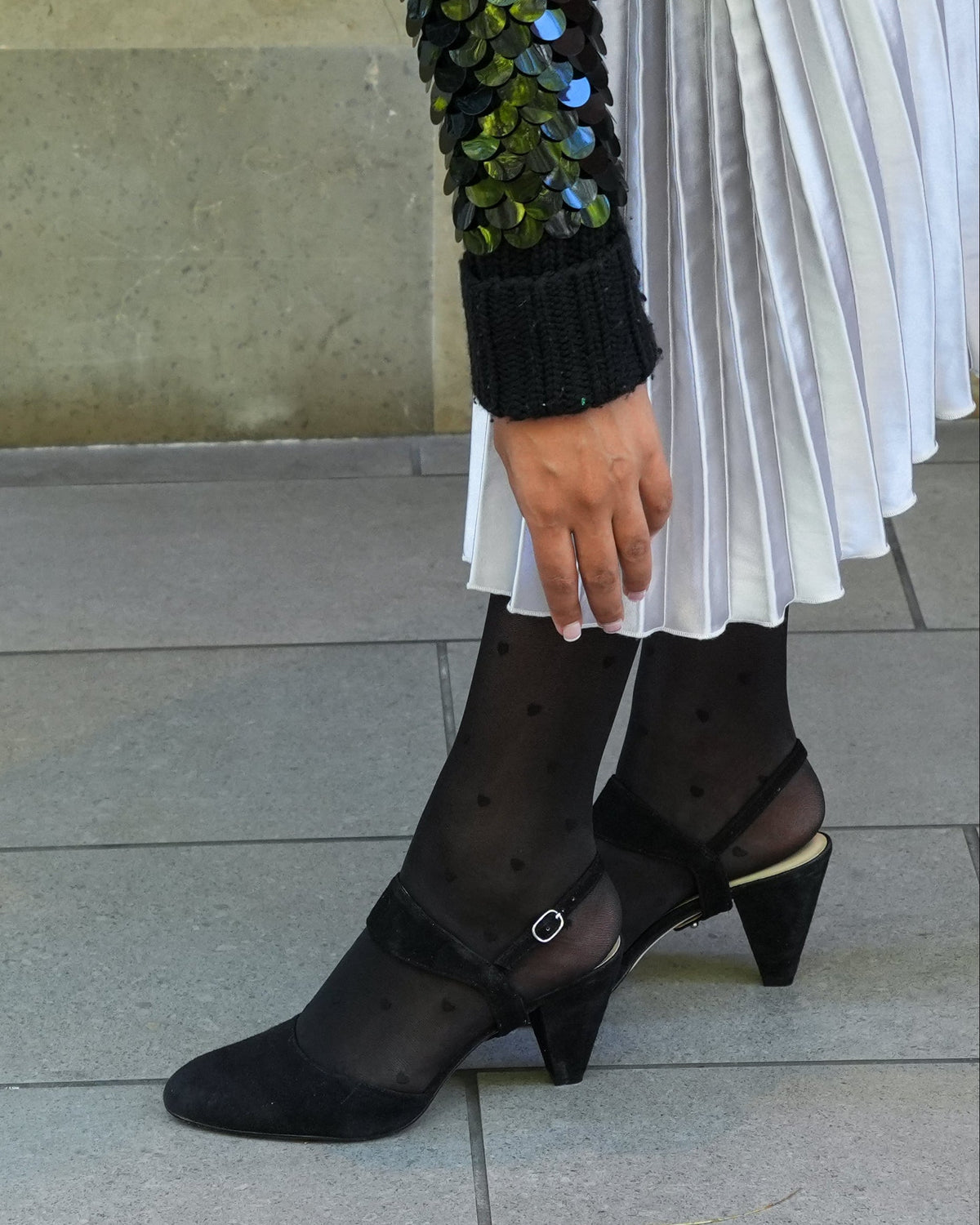 Person wearing black heeled shoes with a patterned sock, standing on a tiled floor.