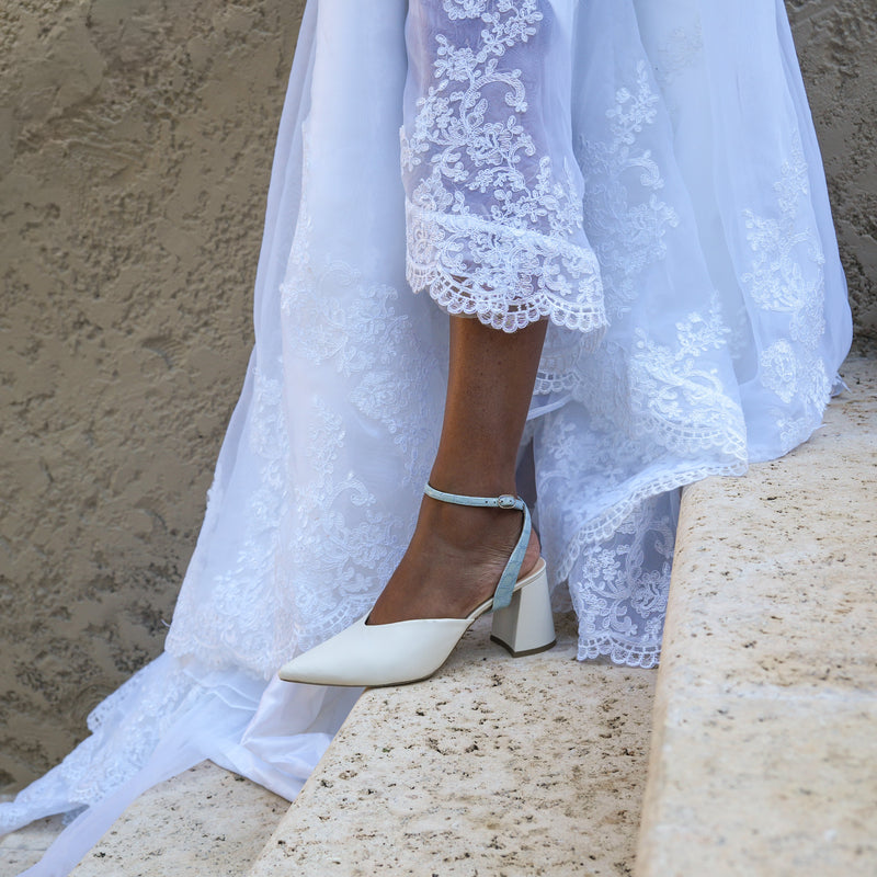 White wedding dress with lace details and cream high-heeled shoes with blue strap on stone steps.