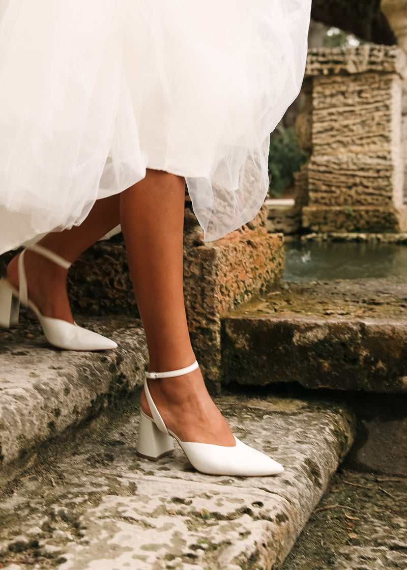 White pointed-toe heeled mule sandals with ankle straps worn by a bride stepping down rustic stone steps beside a fountain.